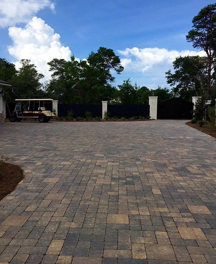A wide paver parking court in mixed tan and gray, running to a dark gate between rendered piers with a golf cart parked at the far side