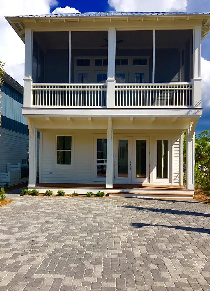 A paver patio at the back of a white house, running up to a screened porch with a white balcony above