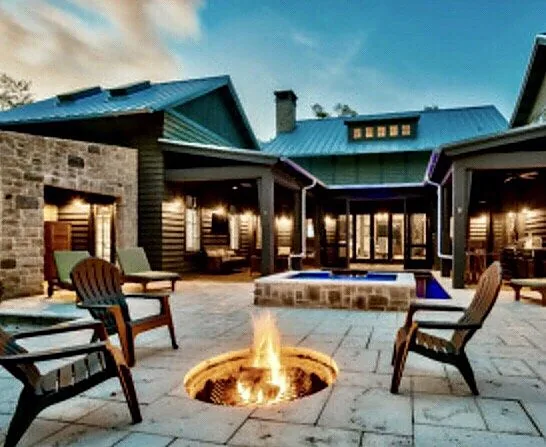 A round fire pit alight at dusk, ringed by wooden Adirondack chairs on a pale paver patio, with a lit house and pool behind