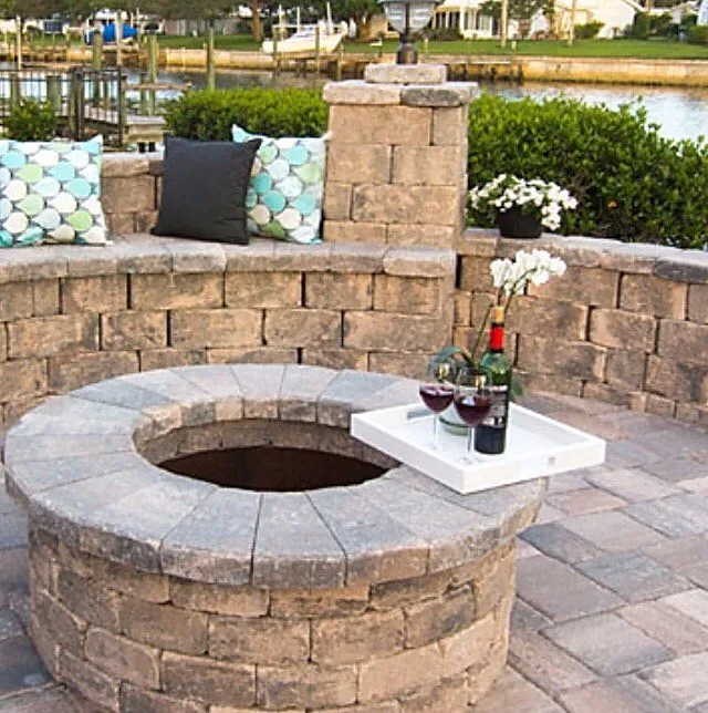 A round stone fire pit with a flat cap on a paver patio, inside a low curved seat wall, with open water behind