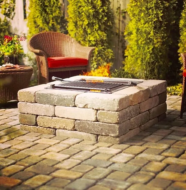 A square stone fire pit alight on a paver patio, with a wicker chair pulled up beside it and planting behind