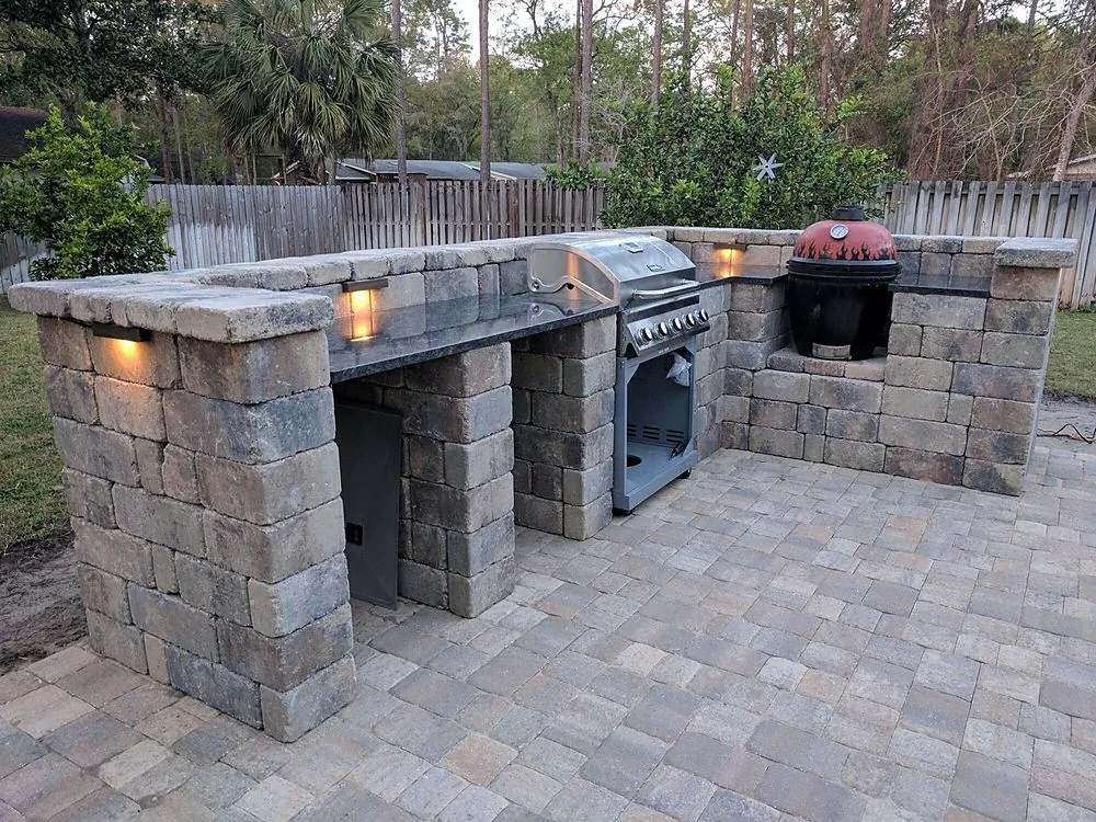 An outdoor kitchen built in gray block with a stone counter and two built-in appliances, its downlights on under flat overcast light