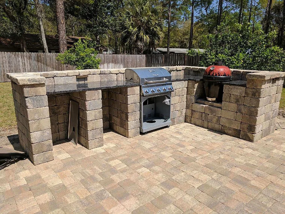 A stone counter run with a red ceramic cooker set into the masonry and a stainless gas grill standing beside it, on a tan paver patio