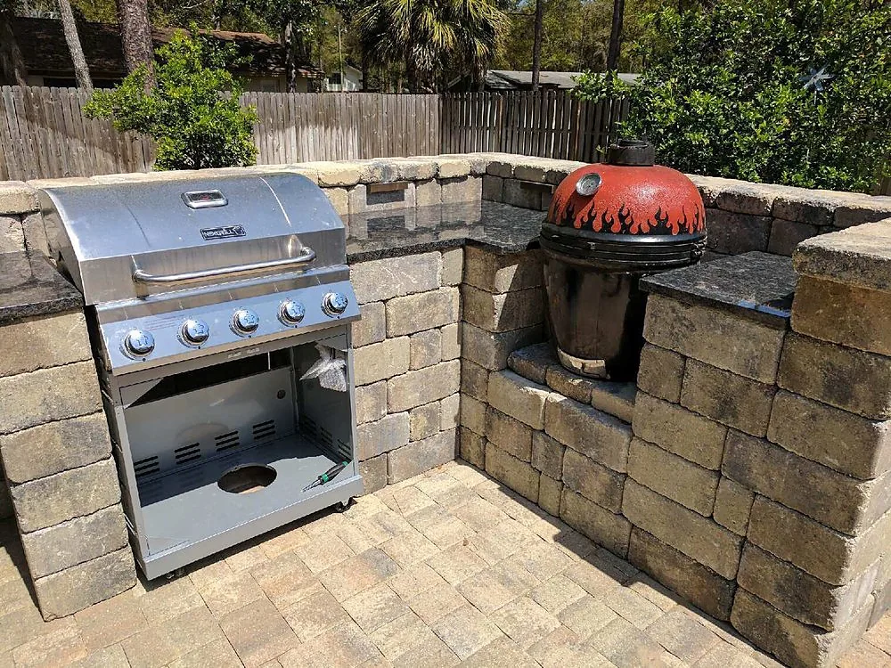 A block seat wall in tan stone, capped flat and wrapping a paver patio, with a red ceramic cooker set into the masonry and a freestanding gas grill beside it