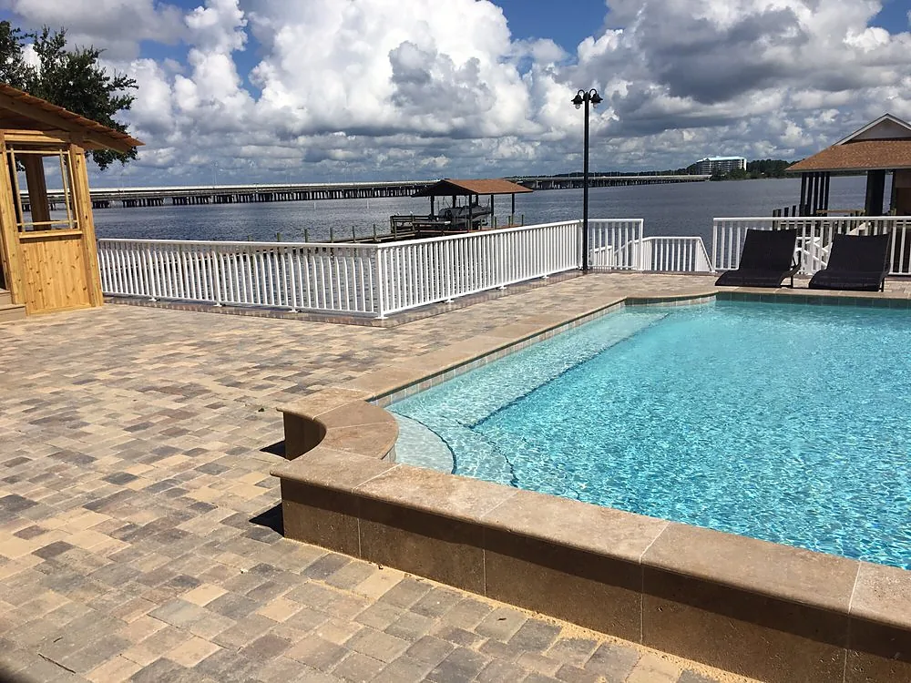 A swimming pool with a paver deck run around it in tan and brown, a low coping course at the waterline and open water beyond the fence