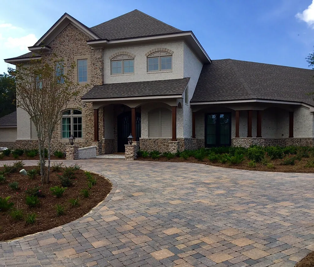 A wide paver driveway running the full frontage of a brick house, with a young tree and a planted bed let into one side