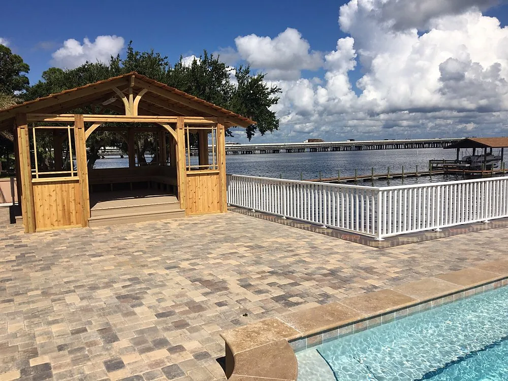 A broad paver deck running out to a white rail and open water, with a roofed cedar pavilion standing on it