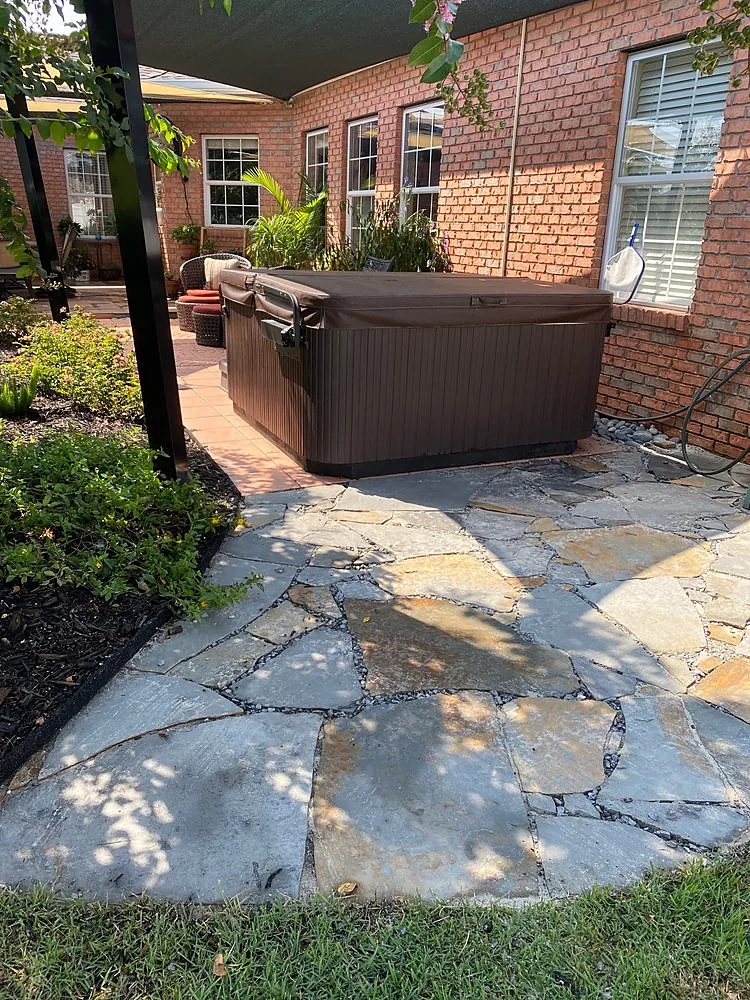 An irregular flagstone patio in gray and cream, the pieces cut and fitted to each other, running through a planted bed to a hot tub