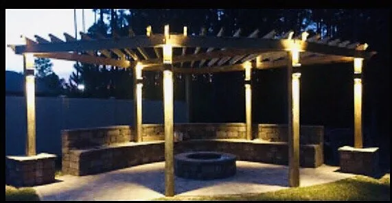 An open-rafter pergola at night, uplit from its timber posts, over a curved seat wall and an unlit stone fire pit