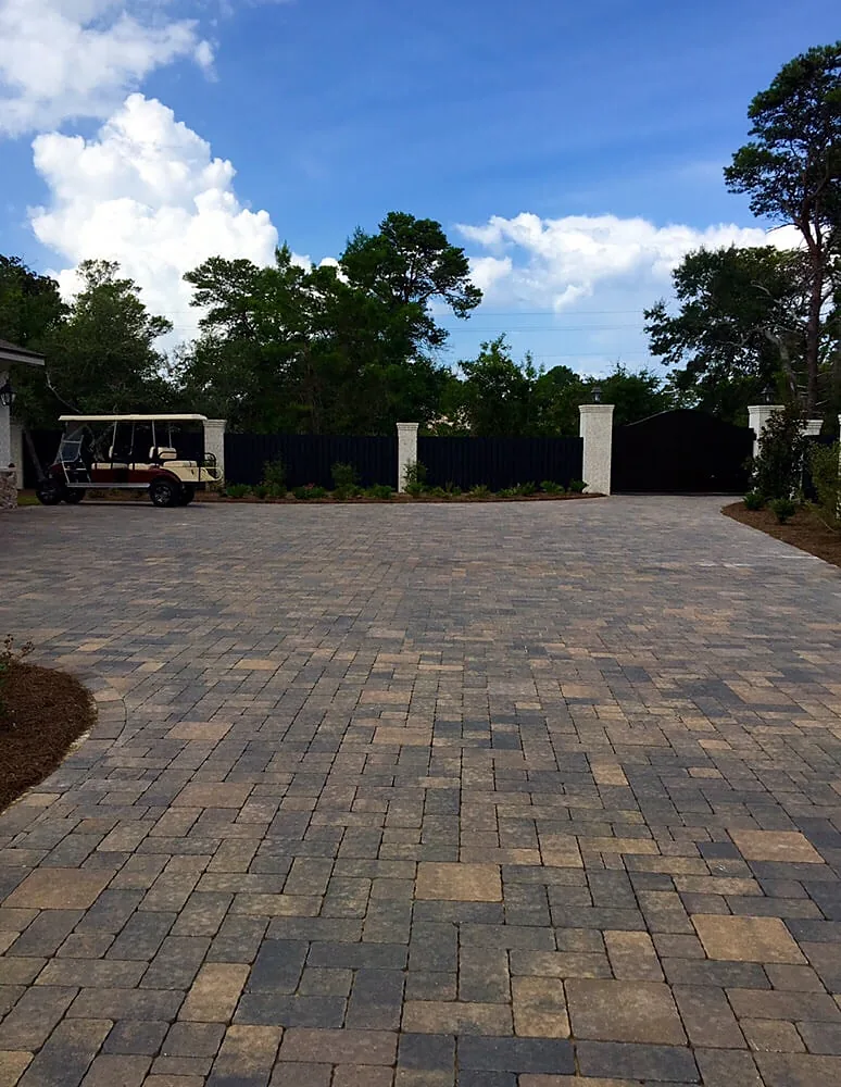 A large paver driveway in mixed tan and gray running the width of the frame toward a house set behind trees