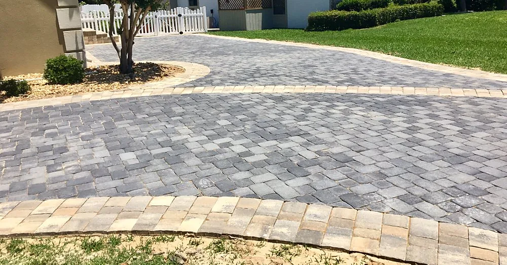 A broad paver driveway in tan and gray curving away past a clipped lawn edge, with a darker border course around it