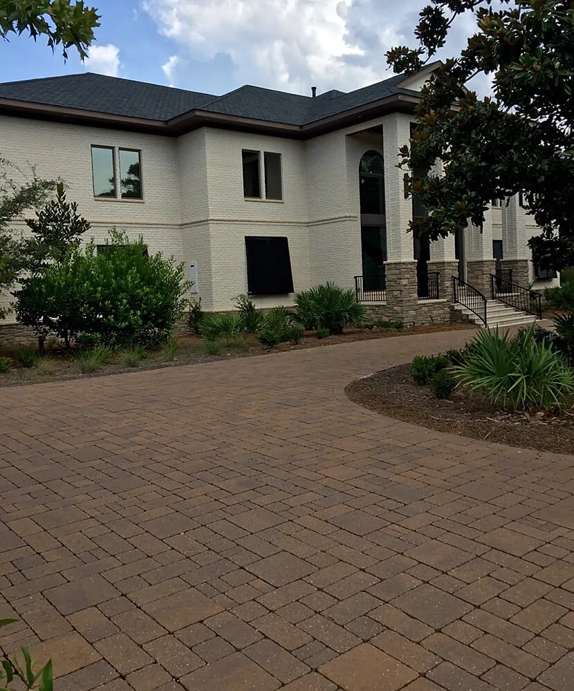 A paver drive and apron in front of a pale stucco house, with planting along one edge