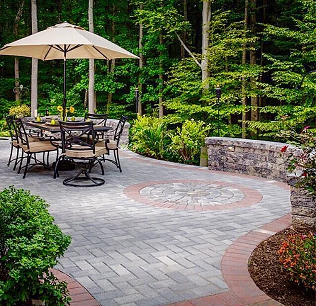 A paver patio in gray with a circular medallion set into the middle and a red border course, with a table, chairs and a cream umbrella, backed by woodland