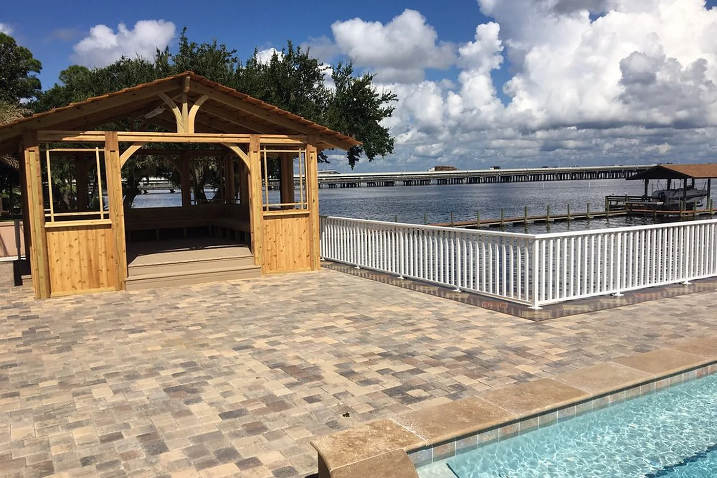 A broad paver deck running out to a white rail and open water, with a roofed cedar pavilion standing on it